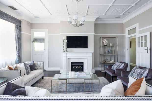 A refined traditional living room with multiple sofas and armchairs arranged around a central glass coffee table and rug. The room features a white fireplace with mantel and mounted television, pale grey walls with white dado rails and trims, ornate decorative ceiling panels, a chandelier, tall drapery, and white framed doors with glazed upper panels.