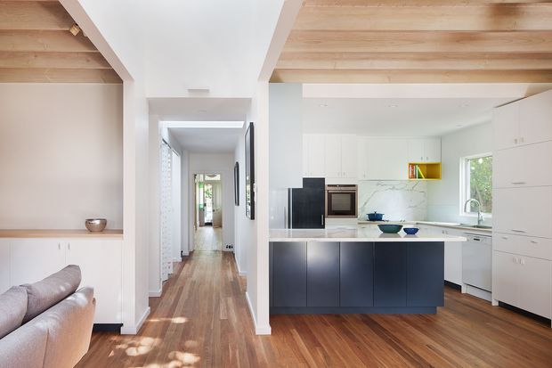 Bright open-plan kitchen viewed from a living area and hallway, featuring a dark island with a light stone benchtop, white full-height cabinetry, integrated appliances, timber flooring, and exposed pale timber ceiling lining. A corridor extends toward the rear of the home, with a small portion of a sofa visible in the foreground.