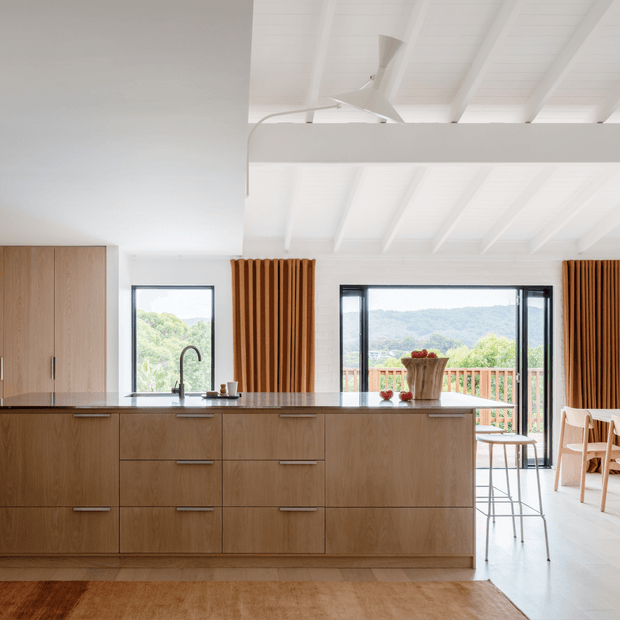 Bright open-plan kitchen with a large timber island, integrated cabinetry, dark stone benchtop, black-framed glazing, and a white vaulted ceiling with exposed beams. The island faces a dining area and balcony overlooking a green landscape. Vertical timber screening and warm wood tones add texture and contrast.