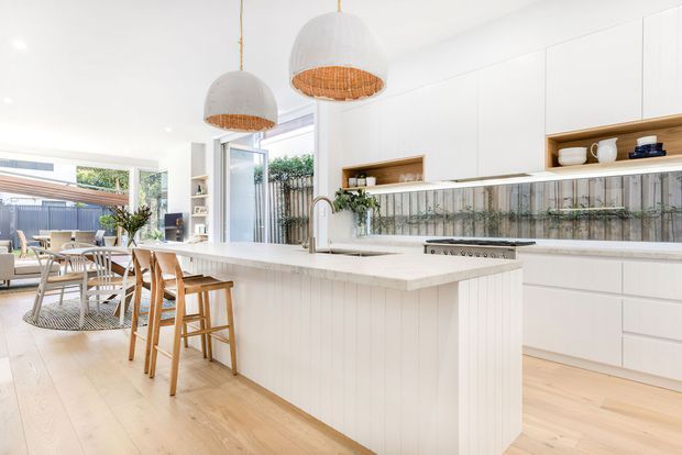 Bright open-plan kitchen and dining area with a large white island, integrated white cabinetry, timber flooring, and a tiled splashback featuring a vertical botanical or textured pattern. The island includes timber bar stools, while woven pendant lights add warmth. A dining and living area opens toward a glazed outdoor connection in the background. The overall design is airy, coastal and contemporary with natural timber accents.