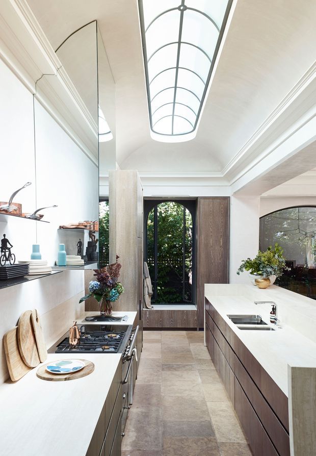 A refined galley-style kitchen with two long runs of cabinetry and pale stone benchtops. Dark timber cabinetry contrasts with light stone surfaces and large-format neutral floor tiles. A dramatic elongated arched skylight brings abundant natural light into the room, while tall arched glazing and a mirrored wall add depth. The kitchen includes a gas cooktop, integrated sink, open display shelving, and decorative floral arrangements.