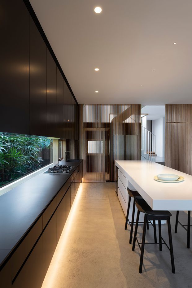 A contemporary galley kitchen with a long dark built-in cabinet and cooking run on the left, a large white island with integrated seating on the right, and warm timber vertical screening at the rear. Recessed ceiling lights, concealed under-cabinet lighting, concrete-look flooring, and extensive glazing create a refined, warm modern interior. Lush planting is visible outside through the window.
