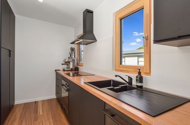 Compact contemporary kitchen with dark flat-panel cabinetry, a warm timber benchtop, integrated black cooktop, deep double sink, black rangehood, and timber-framed window. White walls and a timber-look floor create a clean, warm-toned interior.