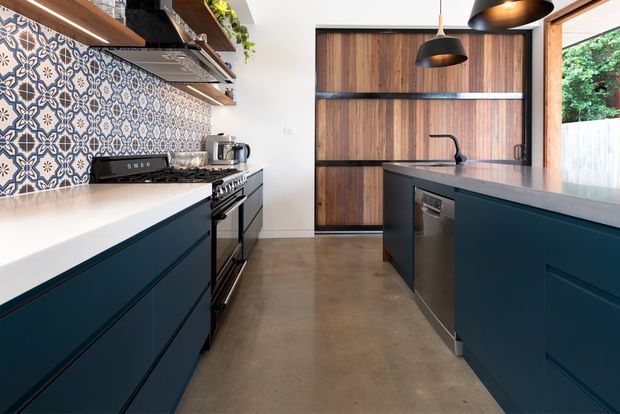A contemporary kitchen with deep blue built-in cabinetry, white benchtops, warm timber wall cabinetry, patterned tile splashback, stainless-steel cooking appliances, black pendant lights, open timber shelving, and a polished concrete floor. The compact galley-style layout includes a central island with sink and dishwasher.