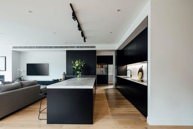 Contemporary open-plan kitchen and living area with a large dark island, black built-in cabinetry, pale stone benchtops, timber flooring, a grey sectional sofa and wall-mounted television. The space has white walls and ceiling, integrated linear lighting, recessed downlights, and a restrained monochromatic palette softened by natural wood and greenery.