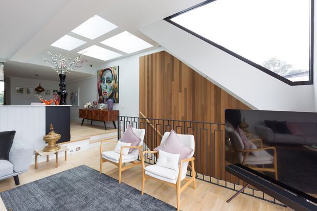 Bright contemporary open-plan living room with a timber feature wall beneath a dramatic sloping ceiling and clerestory-style glazing. The space includes a large television, upholstered armchairs, a grey area rug, a white kitchen island, pendant lighting, and a colourful artwork. A geometric black metal balustrade separates the living area from the adjoining level.