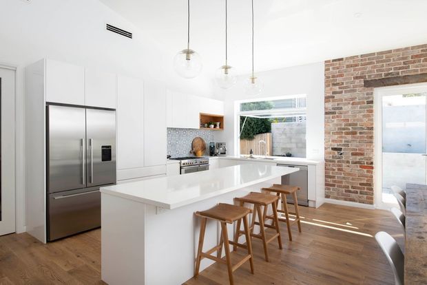 Bright open-plan kitchen with white flat-panel cabinetry, a large white island with timber bar stools, stainless-steel appliances, timber flooring, and exposed brickwork around a glazed opening. Clear pendant lights and generous windows create an airy contemporary interior.
