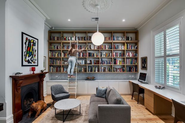 A refined, light-filled living room and home library with a full wall of built-in timber bookshelves and lower white cabinetry. A person stands on a small ladder at the shelving, while a grey sofa, armchair, round coffee table, fireplace, desk, artwork, and large plantation-shuttered windows complete the room. Decorative ceiling moulding and cornices add traditional architectural character, contrasted with contemporary furnishings.