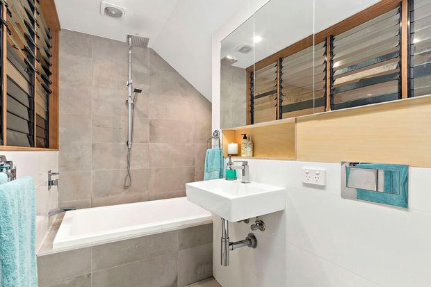 Bright contemporary bathroom featuring a white wall-mounted basin, bathtub with shower, large-format beige-grey wall tiles, white wall finishes, timber-framed louvre windows, mirrored cabinetry or shelving, and stainless-steel plumbing fixtures. Turquoise towels provide a small accent colour.