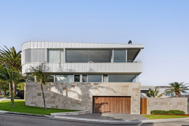Street-facing view of a contemporary multi-level residence with a curved upper floor, extensive glazing, pale vertical metal cladding, a broad cantilevered balcony, and a substantial light stone-clad lower level. A timber garage door, palms, manicured lawn, and a low perimeter wall reinforce the warm coastal setting.