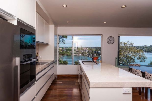 Contemporary kitchen with sleek white full-height cabinetry, integrated stainless-steel appliances, dark timber flooring and a large pale stone island with an undermount sink. Recessed ceiling lights and broad windows provide views over a leafy waterfront setting.