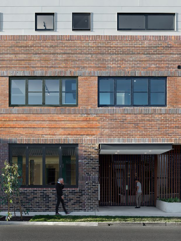 Front-facing view of a contemporary multi-storey brick building facade. The facade combines bands of red and dark brickwork with pale horizontal cladding, dark-framed rectangular windows, glazed ground-floor doors, and a recessed entrance screened by vertical metal slats. A narrow planted strip and sidewalk run along the street frontage, with pedestrians providing scale.
