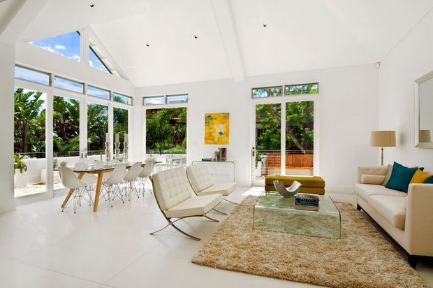 Bright, airy open-plan living room and dining area with a high vaulted white ceiling, extensive floor-to-ceiling glazing, white tiled flooring, and lush greenery visible outside. The room contains a cream sofa, two cream lounge chairs, a glass coffee table, a shag rug, a small dining table with modern white chairs, and colourful cushions and artwork. The overall aesthetic is light, clean, and contemporary with mid-century-inspired furniture.