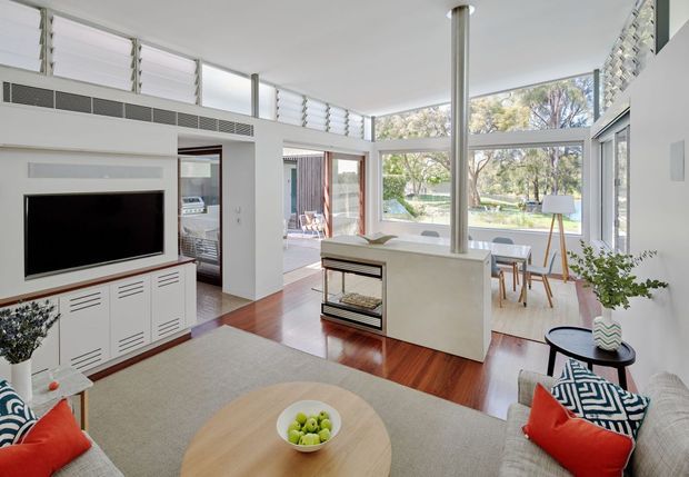 Bright open-plan living room with a contemporary white interior, expansive louvred clerestory windows and floor-to-ceiling glazing overlooking a leafy garden. The room includes a built-in TV cabinet, large pale sectional sofa, timber flooring, a central fireplace or heater enclosure, dining area, and strong indoor-outdoor connection.
