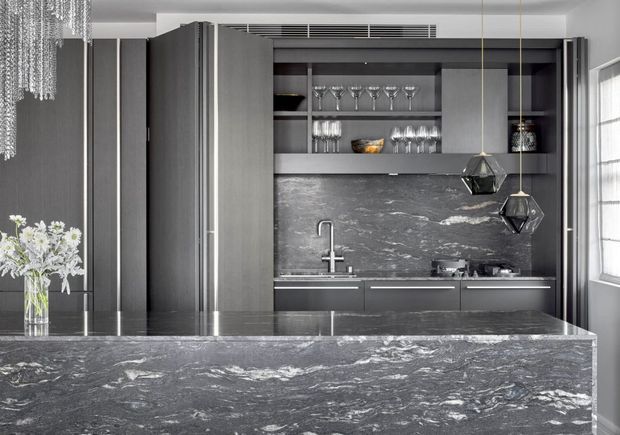 A sophisticated dark-toned kitchen with a large waterfall island and matching stone splashback, integrated dark cabinetry, open glassware shelving, and a compact cooktop and sink zone. Two geometric pendant lights hang over the island, while a crystal chandelier and flowers add decorative detail. A window on the right brings in natural light.