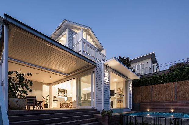 Twilight exterior view of a contemporary multi-level home with pale blue-grey horizontal cladding, extensive glazed openings, balconies, covered outdoor living areas, and broad timber steps leading toward a swimming pool. Warm interior lighting is visible through the glazing, with a neighbouring dwelling and privacy fence in the background.