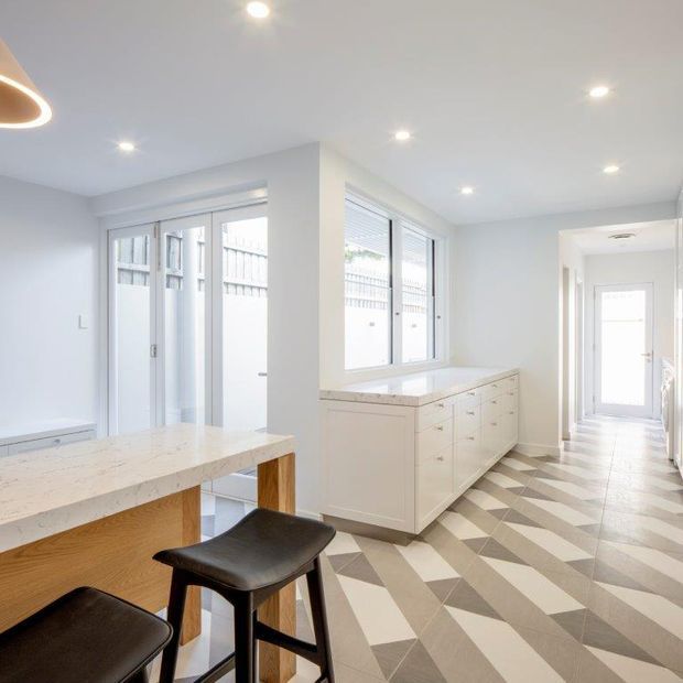 Bright contemporary open-plan kitchen and hallway with white cabinetry, a pale stone island benchtop, timber island joinery, black stools, extensive glazing, and a distinctive geometric patterned tile floor. The space is illuminated by recessed ceiling lights and a sculptural circular pendant.