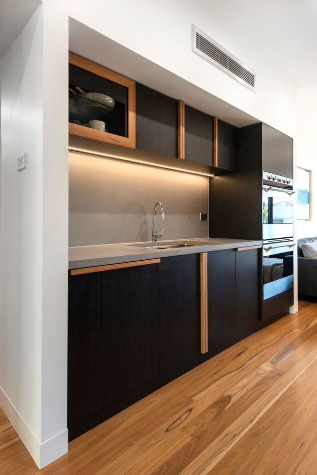A compact contemporary kitchen with dark timber-look cabinetry, warm timber accents, a grey engineered-stone benchtop, integrated stainless-steel sink and tap, and a matching grey splashback. Open shelving with glass-front cabinetry, under-cabinet lighting, integrated oven, and timber flooring create a refined, warm-toned fit-out. A living area is partially visible beyond the kitchen.