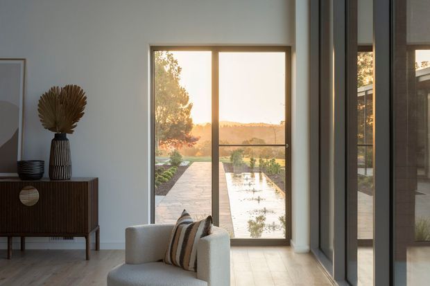 A refined contemporary living room opening onto a landscaped garden and long reflecting pool. The room has pale walls, light timber flooring, full-height dark-framed glazing and a sculptural upholstered armchair beside a dark ribbed timber sideboard with decorative objects. Warm late-day light and the garden view create a calm, elegant atmosphere.
