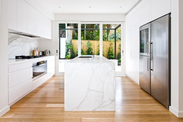 Bright contemporary kitchen with white full-height cabinetry, a large veined white stone island and matching splashback, integrated cooking appliances, stainless-steel refrigerator, timber flooring, and expansive glazed doors opening to a planted outdoor area.