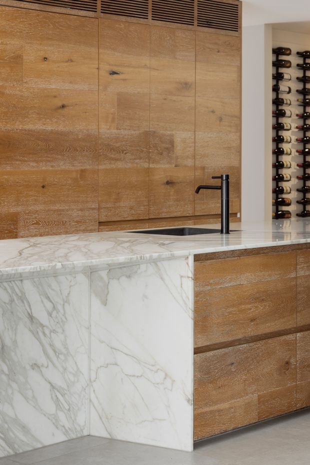 Close-up view of a refined contemporary kitchen centred on a large island. Warm natural timber cabinetry with pronounced grain and integrated ventilation grilles contrasts with a white marble island benchtop and waterfall end panel. A black undermount sink and slim black tap provide contrast. An open wine-bottle storage display is visible in the adjoining wall niche.