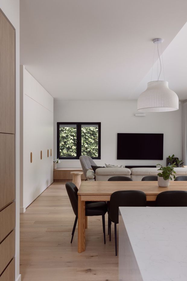 A bright, contemporary open-plan living and dining area viewed from the kitchen. The space features pale timber flooring, white built-in cabinetry with small timber pulls, a timber dining table with dark upholstered chairs, a neutral sectional sofa, a wall-mounted television, and a sculptural ribbed pendant light. A dark-framed window brings in greenery, while a white stone-look kitchen island occupies the foreground.