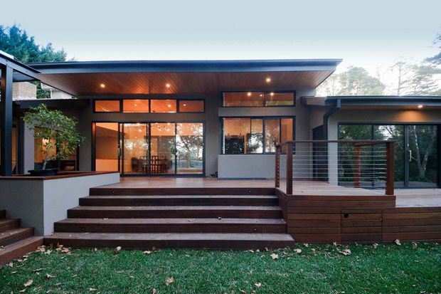 Contemporary single-storey home viewed from the rear garden, featuring broad flat roof planes, dark rendered walls, extensive timber-framed glazing, and a warm timber-lined soffit. Wide timber steps lead up to a raised deck and outdoor living platform with horizontal metal cable-style balustrading. Interior spaces are visible through the glazed doors, while mature trees and lawn surround the house.