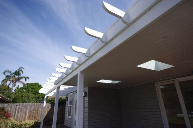 Exterior view of a contemporary single-storey home extension or covered outdoor area, featuring dark horizontal wall cladding, a timber-toned soffit with multiple skylights, slender white support columns, and a distinctive repeating row of curved white roof fins. Tropical planting and a timber boundary fence are visible around the structure.