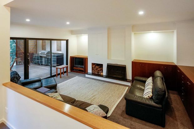 A warm, compact living room viewed from an elevated or open adjoining area. The room features leather sofas and armchairs around a large pale rug, a built-in timber-framed half-wall or balustrade, recessed ceiling lighting, a television in built-in joinery, and a prominent fireplace wall with timber cabinetry. Glazed doors open to an outdoor entertaining area with a barbecue kitchen.