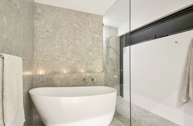 Contemporary bathroom featuring a freestanding white bathtub, large-format pale stone wall and floor finishes, a frameless glass shower enclosure, and minimalist white cabinetry with a dark recessed shelf and towel rail. Recessed lighting creates a calm, refined atmosphere.