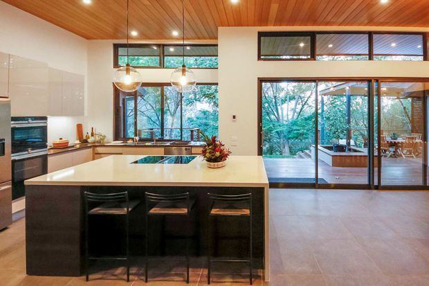 Open-plan kitchen and living area with a large island, dark cabinetry, pale benchtop, bar seating, integrated appliances, warm timber ceiling, and pendant lighting. Full-height glazing and sliding doors open to a covered outdoor entertaining deck surrounded by trees.
