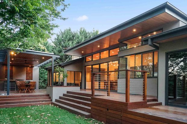 Contemporary single-storey residence viewed from the rear, featuring broad low-pitched roof planes, extensive dark-framed glazing, warm timber soffits, and a raised timber deck with steps and cable-style balustrades. A covered outdoor dining area is visible at left, surrounded by established trees and lawn.