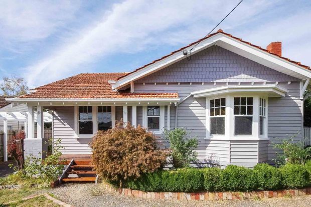 Exterior view of a single-storey character bungalow with pale grey horizontal weatherboard cladding, a terracotta tiled gable roof, decorative gable shingle detailing, white timber trims, and a projecting bay window. A covered front porch, timber steps, clipped hedges, and established garden landscaping complete the street-facing facade.