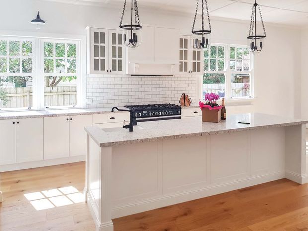 Bright, spacious traditional-style kitchen with a large central island, white shaker cabinetry, glass-front upper cabinets, subway-tile splashback, stone benchtops, timber flooring, and multiple pendant lights. Large windows provide abundant natural light and views of greenery outside.