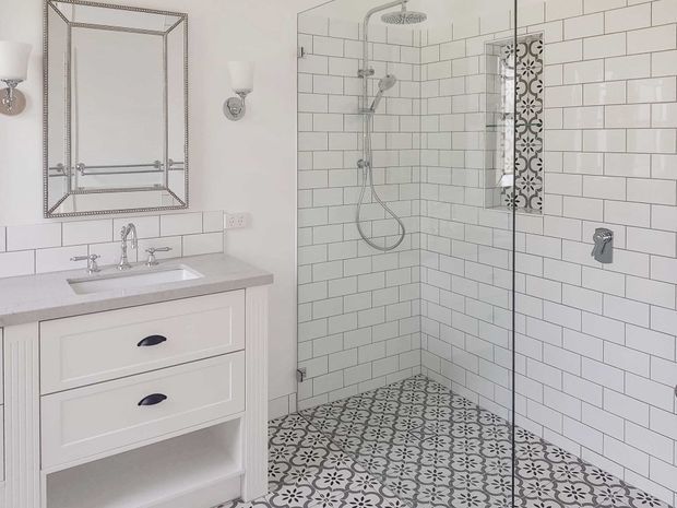 Bright traditional bathroom with a white shaker-style vanity, pale stone benchtop, chrome fixtures, framed mirror, subway-tiled walls, and a large walk-in shower enclosed by frameless glass. The shower floor features patterned encaustic-style tiles, with a matching decorative vertical tile accent in the wall niche.