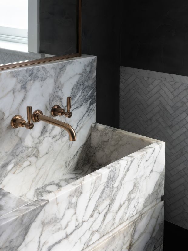 Close-up of a luxurious bathroom washbasin area featuring a sculptural white-and-grey veined marble basin and matching marble splashback. Brushed brass wall-mounted tapware contrasts with dark charcoal wall surfaces, while a grey marble herringbone tile wainscot runs along the adjacent wall. A timber-framed mirror and window are visible above the vanity.