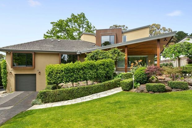 A landscaped suburban residential exterior featuring a low-pitched tiled roof, rendered walls, extensive clipped hedging, and a contemporary timber-clad entry or porch addition with a broad sloping roof. A garage and driveway are visible at the left, with a manicured lawn and layered garden beds in the foreground.
