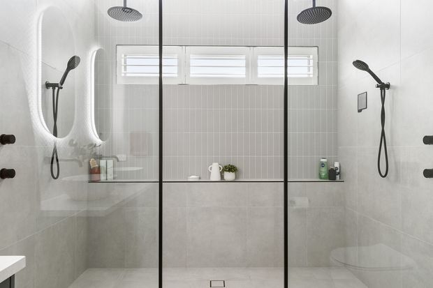 Bright contemporary walk-in bathroom with a large frameless-style black-framed glass shower enclosure, dual ceiling-mounted rain showers and wall handsets, pale grey tiled walls, a concrete-look lower wall finish, and high horizontal windows. Minimal fixtures and recessed shelving create a clean, modern appearance.