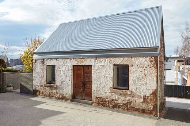 A small, weathered masonry building with rough rendered stone or concrete walls, dark timber-framed windows, a wooden entry door, and a steep corrugated metal gable roof. It sits in a compact paved urban setting with neighbouring buildings visible behind it.