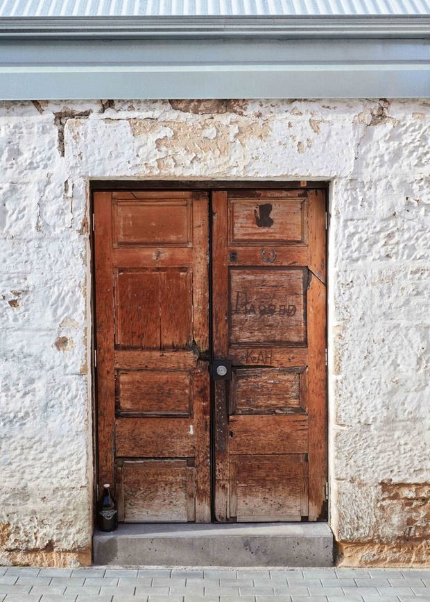 Close-up exterior view of an aged, weathered timber double door set within a rough white-rendered masonry wall. The door has recessed panels, visible patina, graffiti, dark metal hardware, and a simple concrete threshold beneath a corrugated metal roof edge.