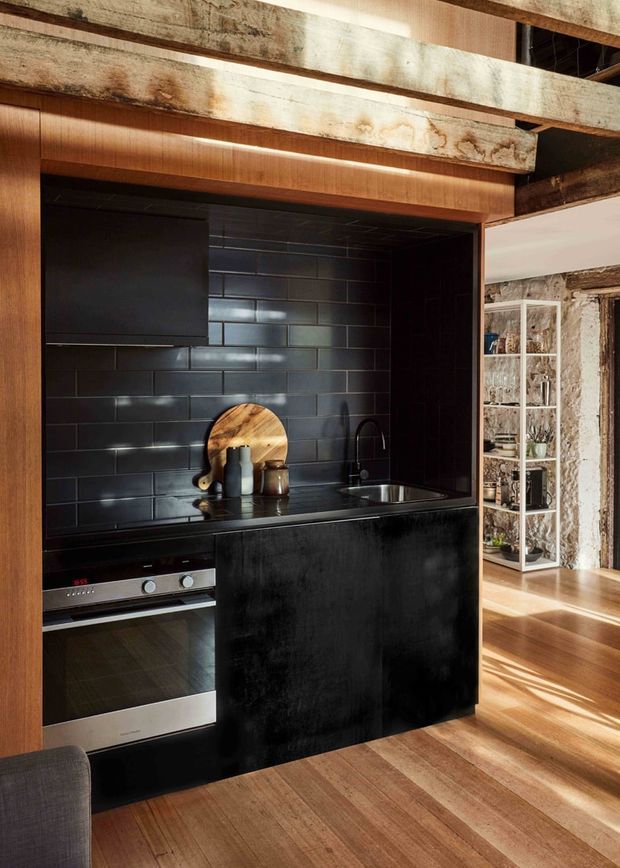 Compact contemporary kitchen with a black tiled splashback, black island cabinetry and integrated stainless-steel oven and cooktop. Warm timber cabinetry, flooring and exposed structural beams contrast with the dark finishes. An open shelving unit and adjoining timber-floored space are visible beyond the kitchen.
