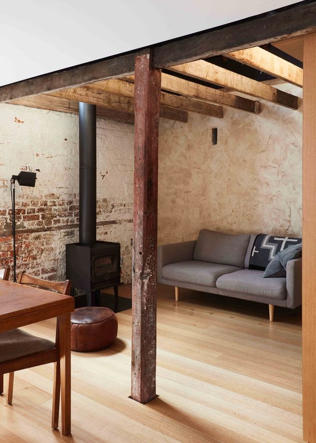 A warm industrial-style living room in a converted or renovated brick structure, featuring exposed timber beams and a central timber post, distressed exposed brick and plaster walls, pale timber flooring, a black wood-burning stove, and a mid-century-inspired grey sofa. A timber dining table and chairs occupy the foreground, with a small brown leather ottoman nearby.