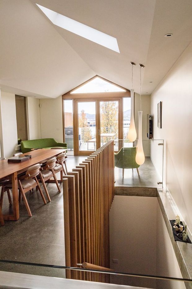 Bright, contemporary open-plan interior viewed from an upper stair landing. The space combines a dining area with a long timber table and upholstered chairs, a green sofa and lounge chair, polished concrete or dark stone flooring, and a timber slatted stair balustrade. Floor-to-ceiling glazed doors open to a balcony with distant landscape views. A prominent triangular clerestory window and angular skylight bring daylight into the high, sloping ceiling, while sculptural pendant lights hang over the stair and seating area.