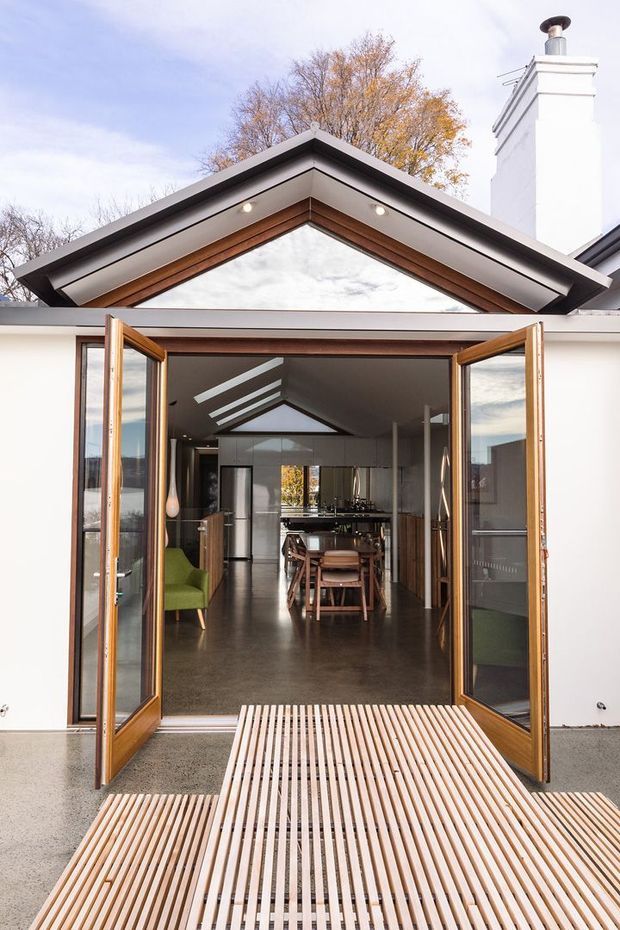 Front-facing view of a contemporary house extension with wide-open timber-framed glazed doors leading into a combined kitchen and dining area. The entry has a pitched gable roof with timber soffit detailing, white exterior walls, a tall white chimney, polished concrete flooring, and a slatted timber deck or bridge in the foreground. Inside are a dining table, kitchen cabinetry, pendant lighting, and a green armchair.