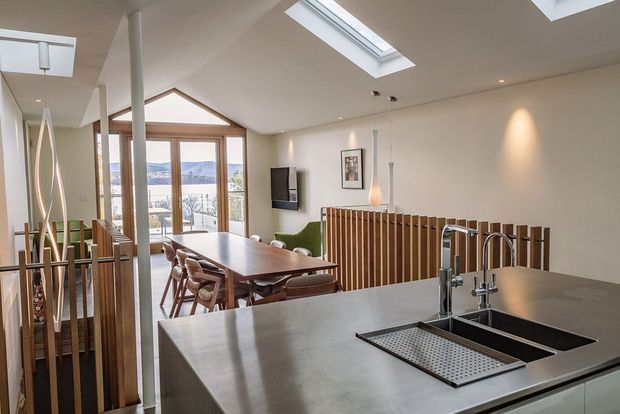 Open-plan contemporary kitchen and dining area with a large stainless-steel island, integrated double sink and drainer, timber dining table, pendant lighting, timber slat balustrade, skylights, and full-height glazing opening to a balcony or exterior view. The space is bright and connected to the dining and living areas.