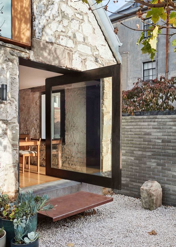 Exterior view of a characterful stone building with a large dark-framed glazed pivot door opening onto a small gravel courtyard. The facade combines weathered pale masonry, timber-framed glazing and dark metal framing, with a raised rust-coloured entry slab, retaining wall, sculptural stone and potted planting. A dining area is visible through the open doorway.