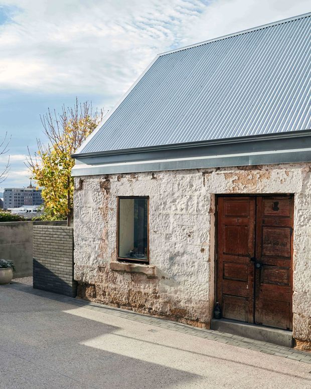 Exterior view of a compact heritage stone building with rough white-painted masonry walls, a steep corrugated metal gable roof, and a dark timber double door. The weathered facade sits beside a paved outdoor area and low masonry boundary wall, with urban buildings visible in the background.