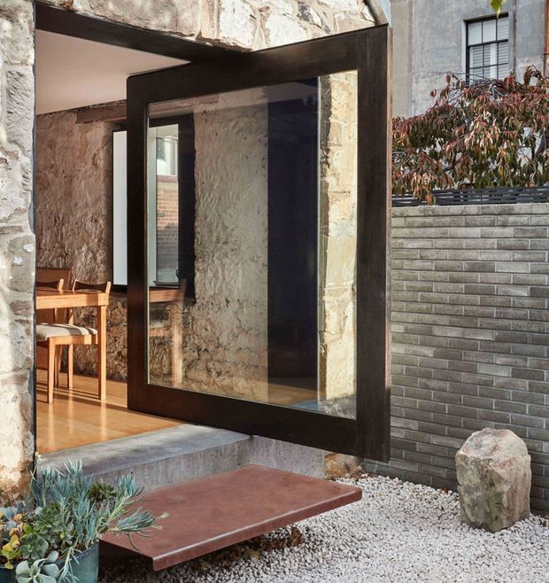 Architectural exterior view of a contemporary entry composed of a large dark-framed glazed pivot door set into a heavily textured stone or masonry wall. The entry opens onto a compact landscaped courtyard with concrete steps, a reddish paved landing, white gravel, succulents, and a dark grey brick boundary wall. A timber-furnished interior is visible through the doorway, with an upper-level window and planting beyond.