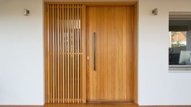 Front entry facade featuring a tall warm timber pivot door with a long dark vertical pull handle, paired with a matching vertical timber-slat security or privacy screen. The entry is set within a smooth white rendered wall, with compact wall-mounted exterior lights and a narrow adjacent window revealing part of the interior.
