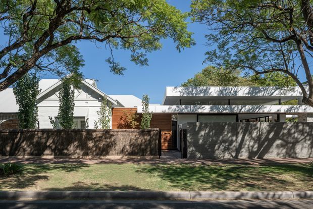 Street-facing view of a contemporary residential property behind a low masonry boundary wall, with a white gabled house visible to the left and a newer flat-roofed white extension or main volume to the right. Timber and brick elements, dark metal-framed openings, mature street trees, and a grass verge are visible under clear daylight.
